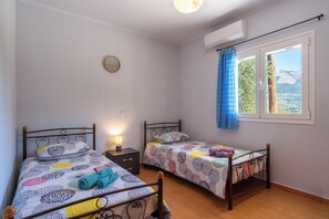 A bedroom with two single beds, a bedside table with a lamp, a wall clock, and a window with a view of the outdoors. - Effie's Escape Spacious House in Zola (Argostoli)