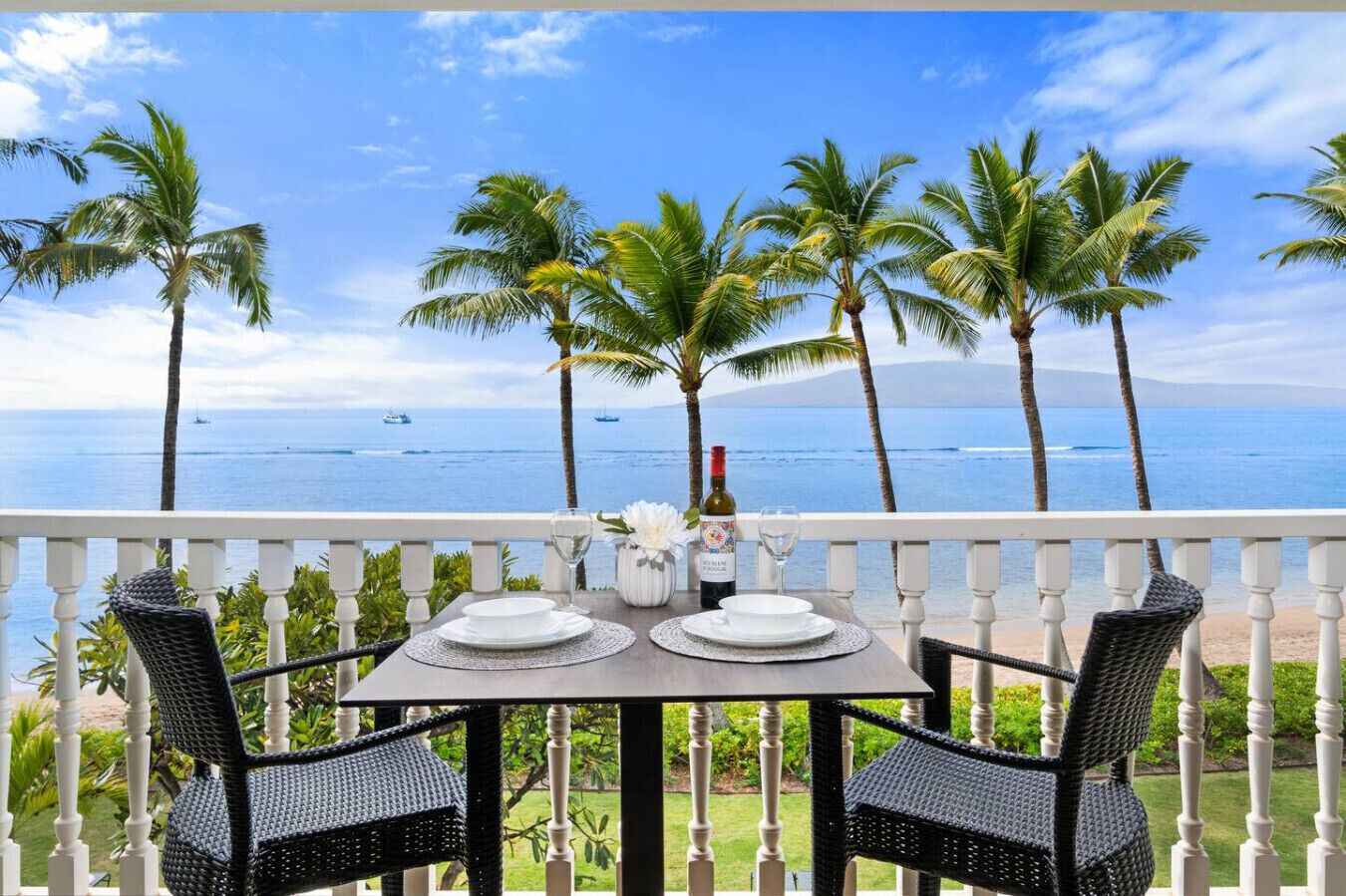 A balcony with a table set for two, overlooking a beach with palm trees and the ocean.