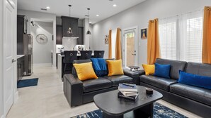A modern living room with a black leather sofa, a coffee table, and a dining area with a kitchen in the background. - Smart 3BR Home + Luxe Decor + Bluetooth Audio (Houston)