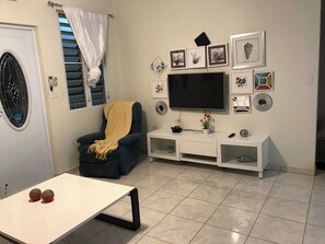 A living room with a TV, wall art, a chair with a blanket, and a small table. - 2-bedroom ocean view villa in lovely Boquerón with AC and charm (cabo rojo)