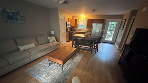 A living room with a sofa, coffee table, and TV. There is a kitchen in the background with a dining table and chairs. - •	Quiet 3BR Home Close to Biddeford Beaches (Biddeford)