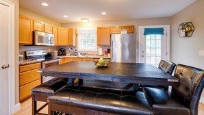 A modern kitchen with wooden cabinets, a stainless steel refrigerator, a large island with a fruit bowl, and a dining area with leather chairs. - •	Quiet 3BR Home Close to Biddeford Beaches (Biddeford)
