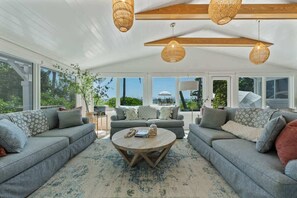 Ein geräumiges Wohnzimmer mit einem großen runden Couchtisch, grauen Sofas und Lampen mit Korbhüllen an der Decke.