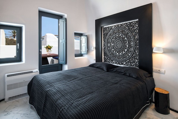 A bedroom with a bed, a door, a window, a wall-mounted decorative panel, and a bedside table with a lamp. - Mirinda Villa (Santorini)