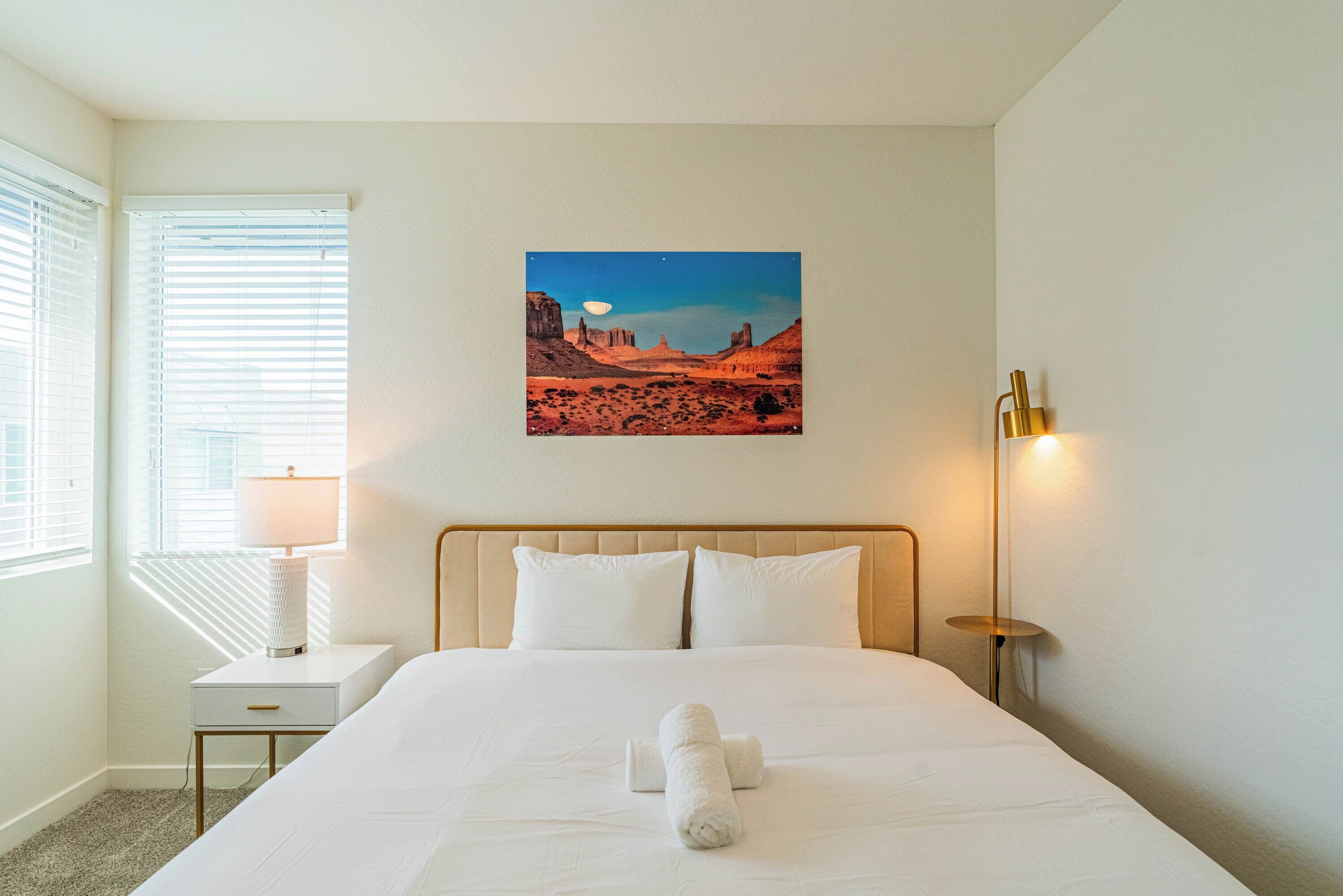 A neatly made bed with white linens, a bedside lamp, a small bedside table, and a wall-mounted picture of a desert landscape.