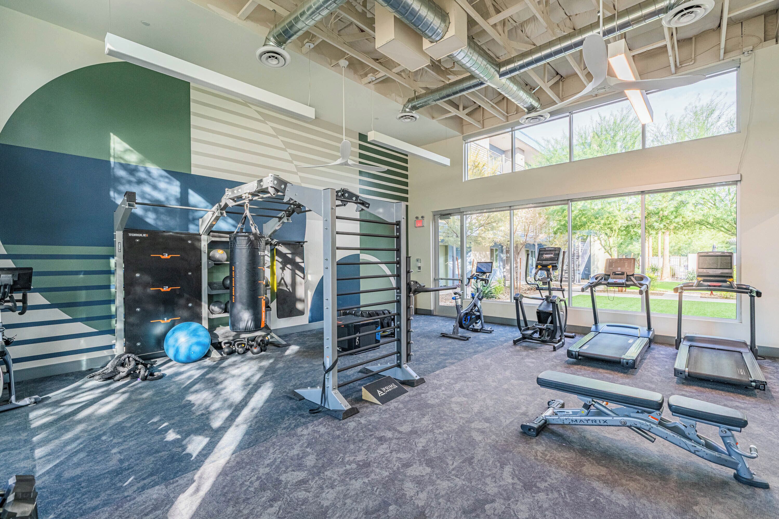 Ein Fitnessstudio mit Trainingsgeräten, Laufbändern und einem großen Fenster mit Blick ins Freie.