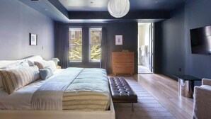 Une chambre moderne avec un grand lit, une commode en bois, un banc et un téléviseur fixé au mur.