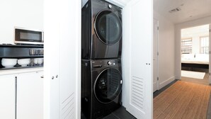A laundry room with two stacked front-loading washers and dryers, a microwave above the sink, and a bed visible through an open door.