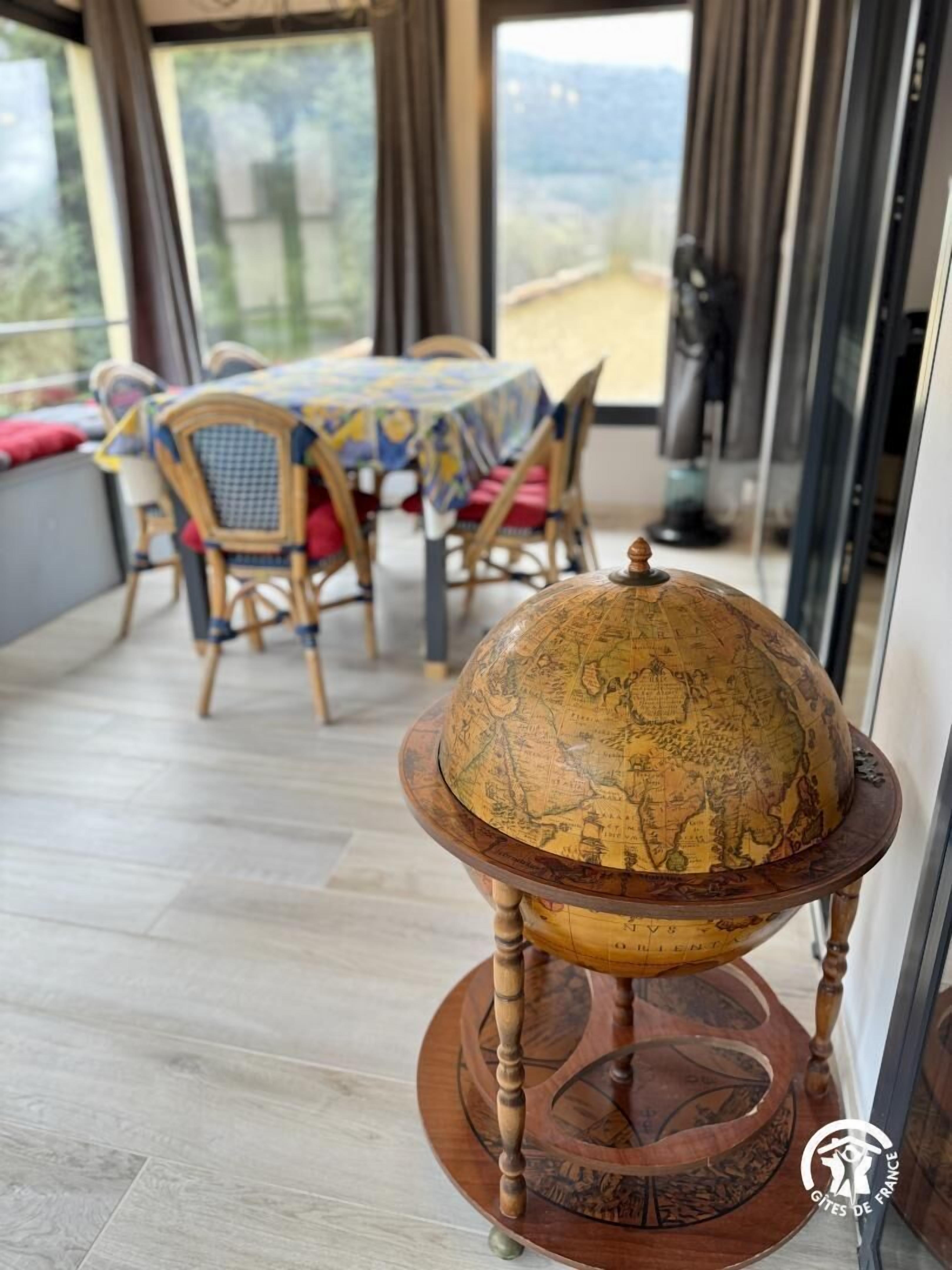 Un globo di legno su un supporto in una stanza con tavolo e sedie, ampie finestre e vista sull'esterno.