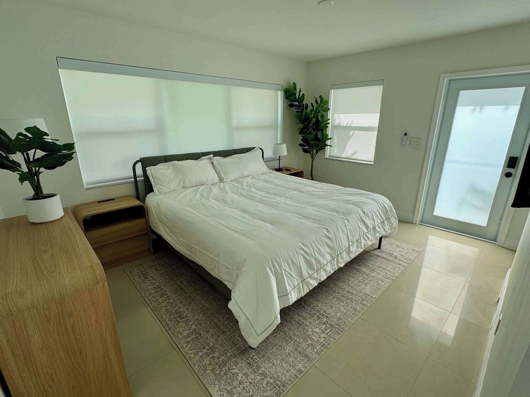 Une chambre moderne avec un grand lit, des tables de chevet, une petite table, une plante en pot et une porte-fenêtre coulissante.