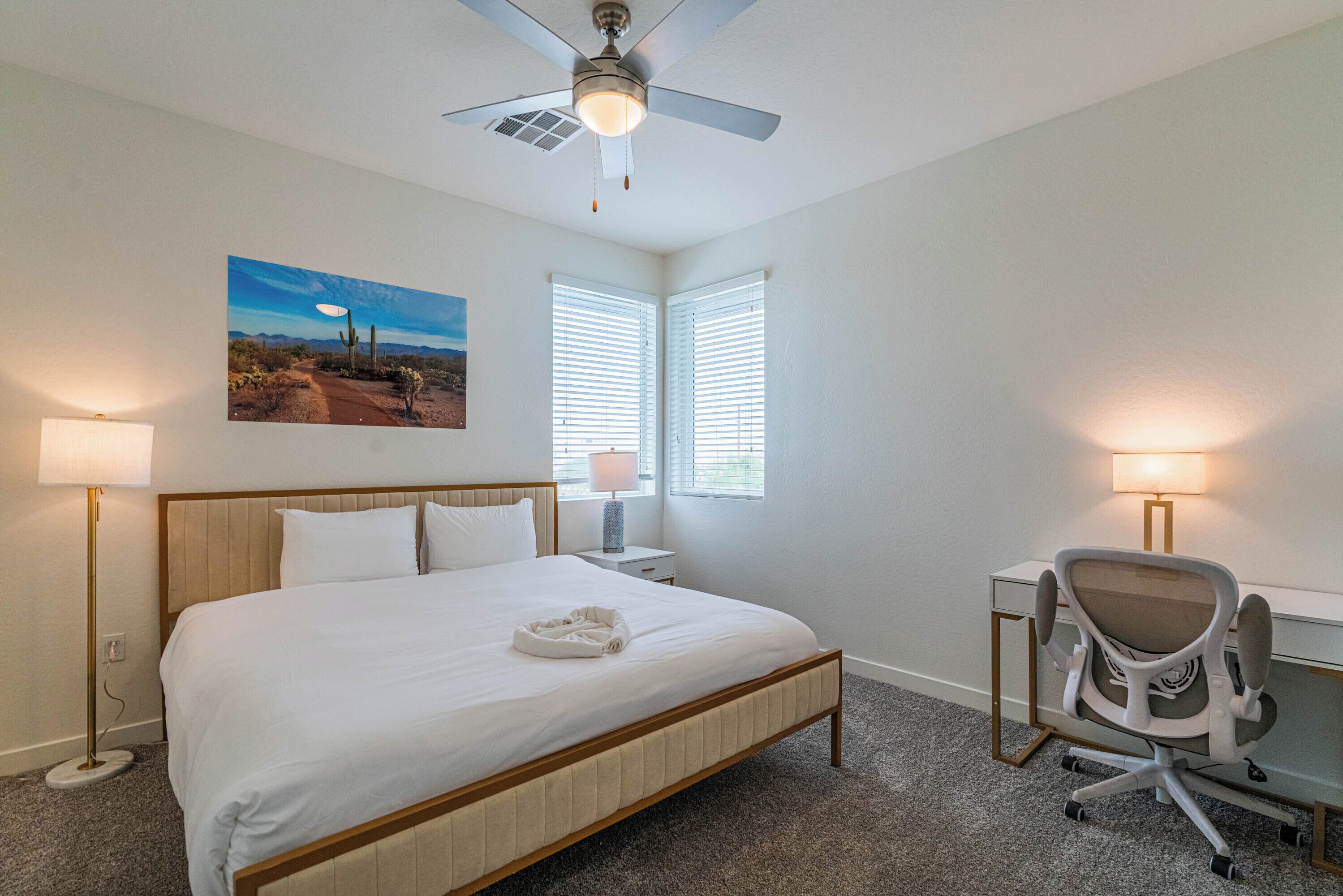 A hotel room with a large bed, a desk with a chair, two windows with blinds, a ceiling fan, and a painting on the wall.