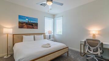 A hotel room with a large bed, a desk with a chair, two windows with blinds, a ceiling fan, and a painting on the wall.