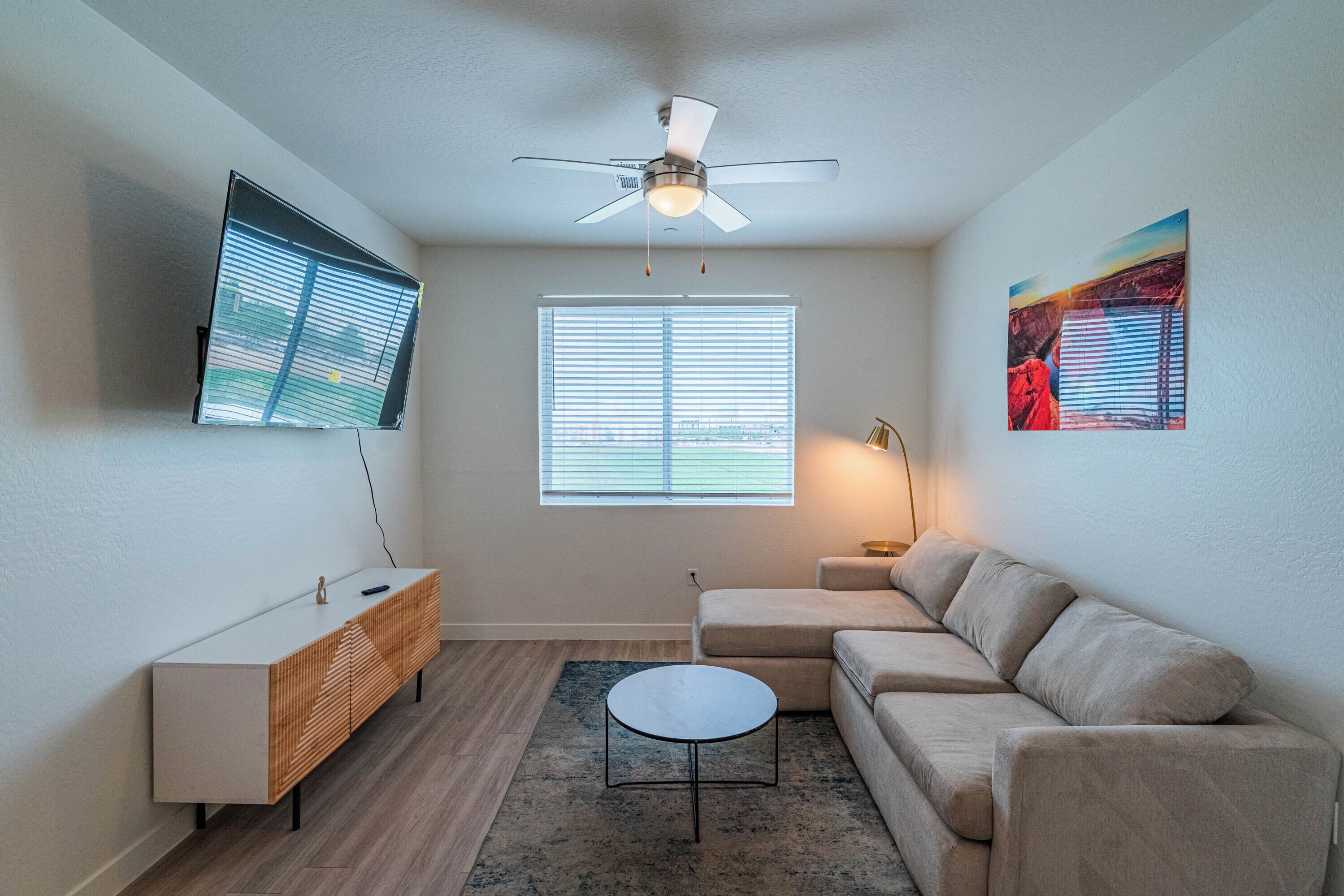 A modern living room with a ceiling fan, a flat-screen TV, a sofa, a coffee table, and a window with blinds.
