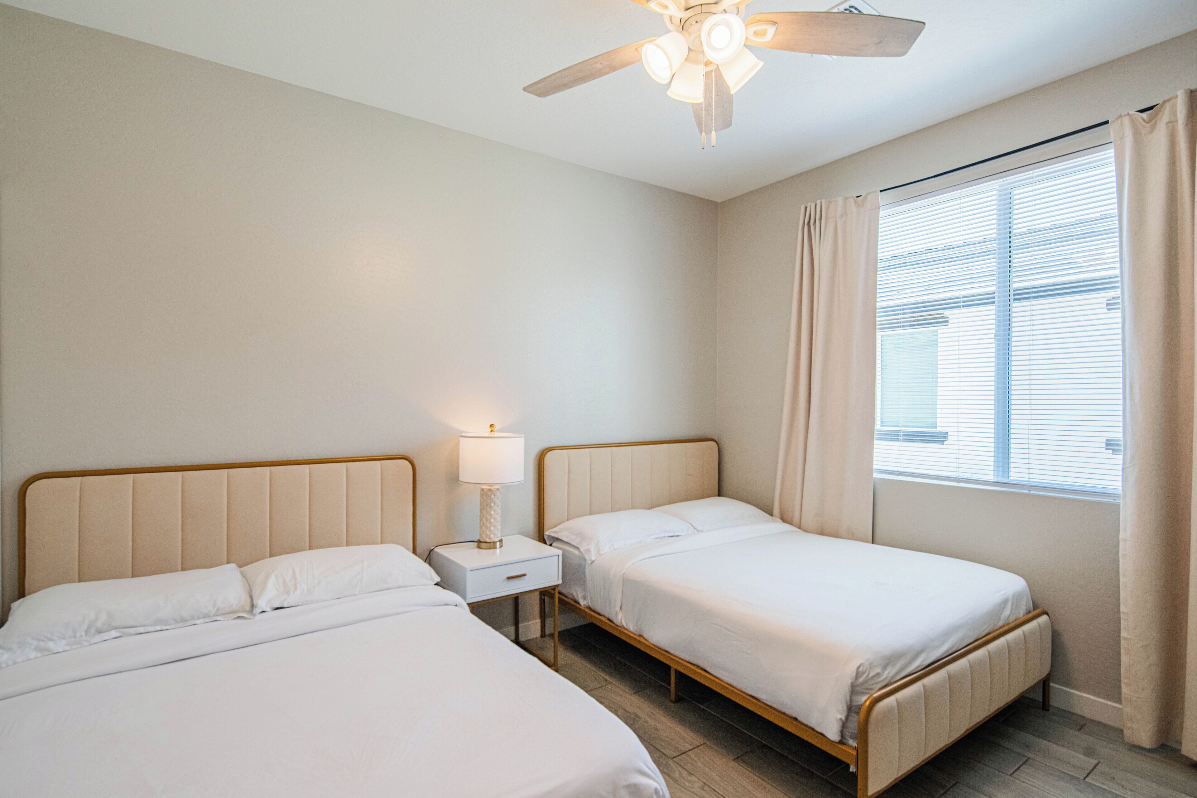 A bedroom with two beds, a ceiling fan, a window with curtains, and a nightstand with a lamp.