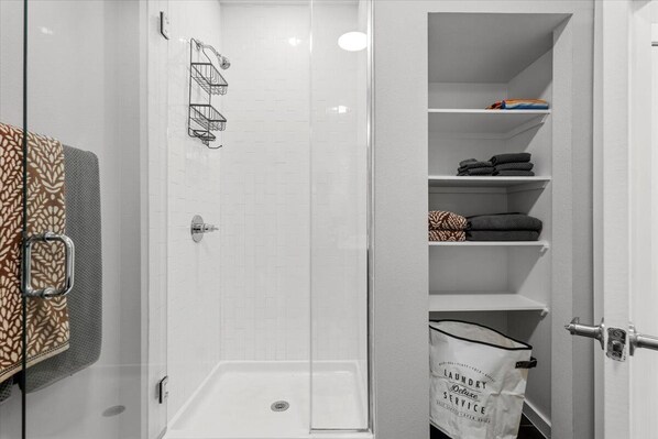 Un closet amplio con paredes de azulejos blancos, un box de ducha de vidrio, y repisas con toallas dobladas y una bolsa para la ropa.