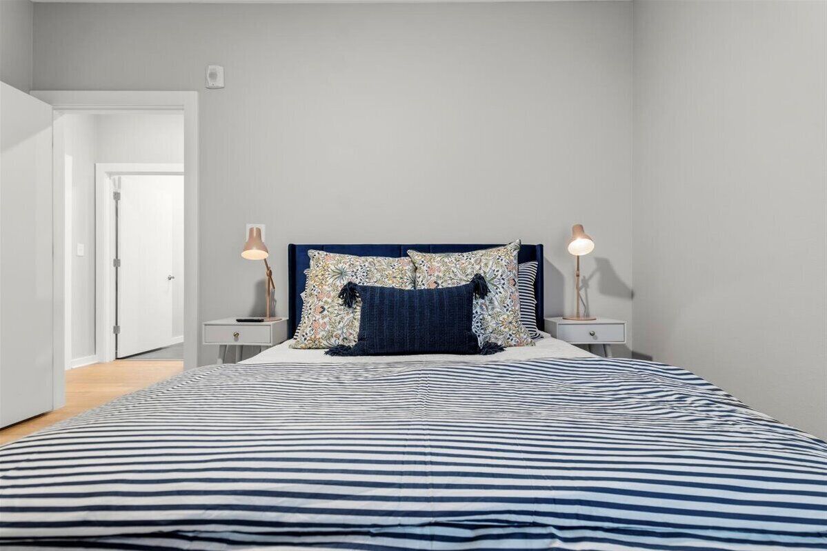 A bedroom with a striped bedspread, a headboard with floral pillows, two bedside lamps, and a white door leading to another room.