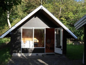 A small cabin with a sliding glass door, a porch with a chair, and a view of the surrounding forest. - Nature oasis with private lake near beach (Fårevejle)