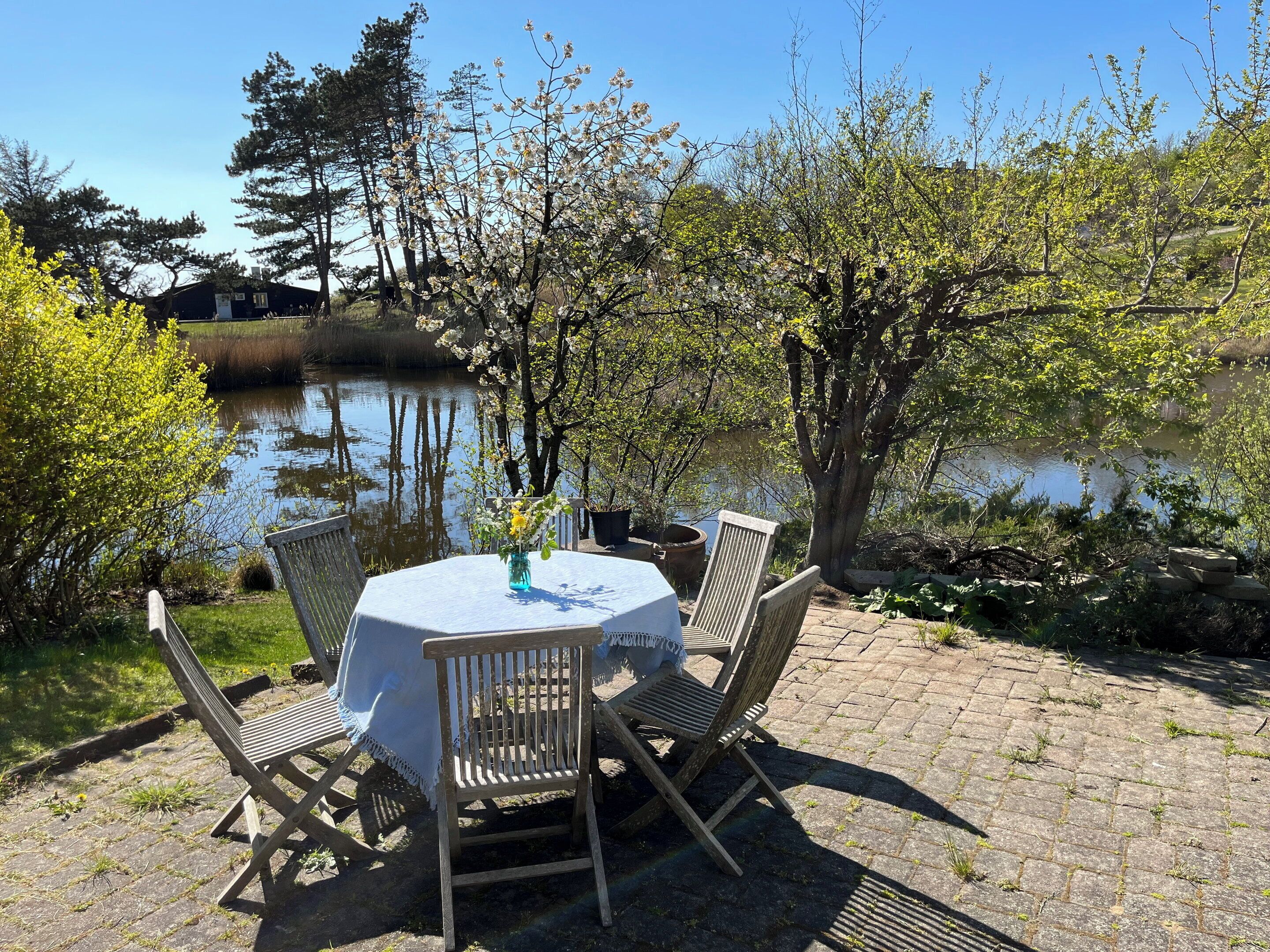 Een ronde tafel met een witte tafelkleed, omringd door houten stoelen, op een terrras van bakstenen met uitzicht op een meer en bomen.