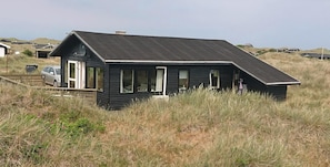 A small, dark wooden house with a sloped roof, a garage, and a car parked outside. - Welcome to this house in the dunes. (Løkken)
