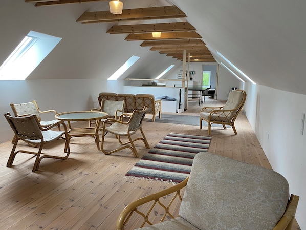 A spacious loft with a high ceiling, wooden beams, a dining area with wicker furniture, and a view of the outdoors through skylights.