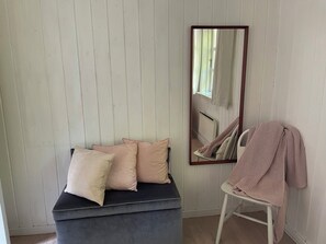 Une pièce avec un banc de couleur sombre, une chaise blanche recouverte d’une couverture rose et un grand miroir accroché au mur.