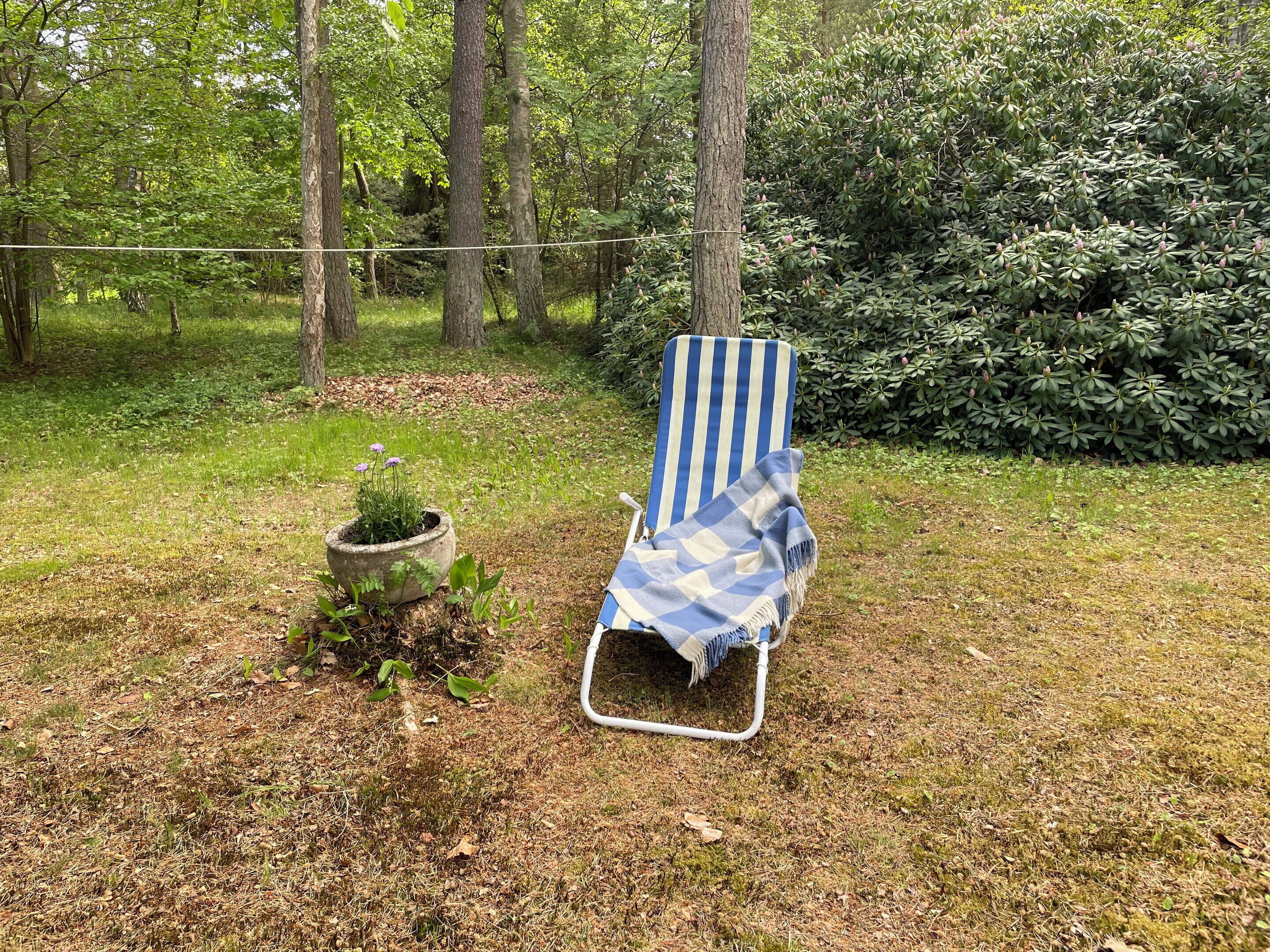 A single lounge chair with a striped backrest in a grassy outdoor area with trees and a hedge.