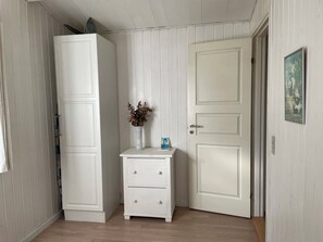 Une pièce avec une armoire blanche, une petite commode blanche avec des tiroirs, une porte avec une poignée et un tableau encadré accroché au mur.