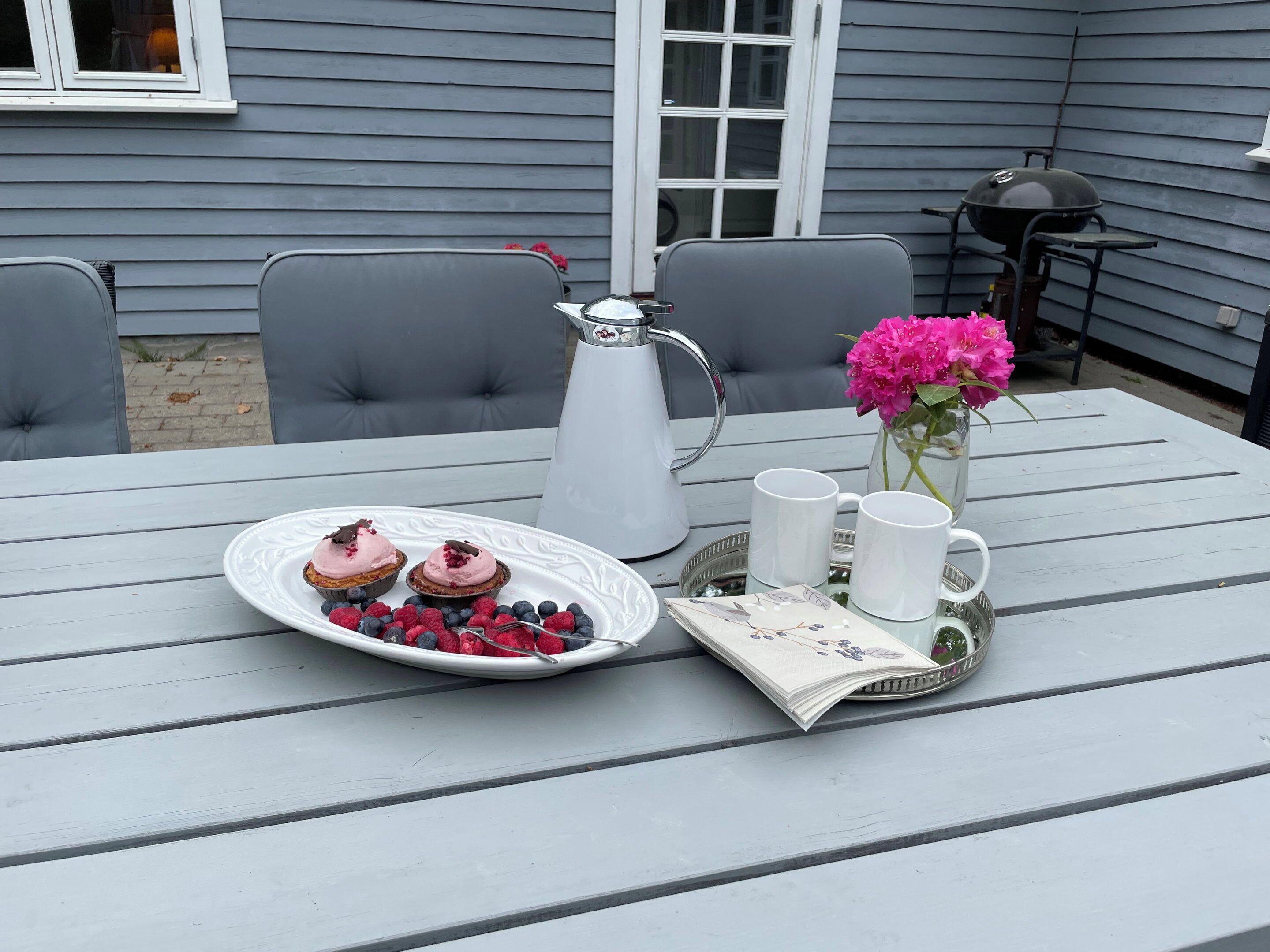 A table with a white plate of desserts, a pitcher, two white mugs, and a vase with flowers.