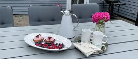 Una mesa con un plato blanco de postres, una jarrilla, dos tazas blancas y un florero con flores.