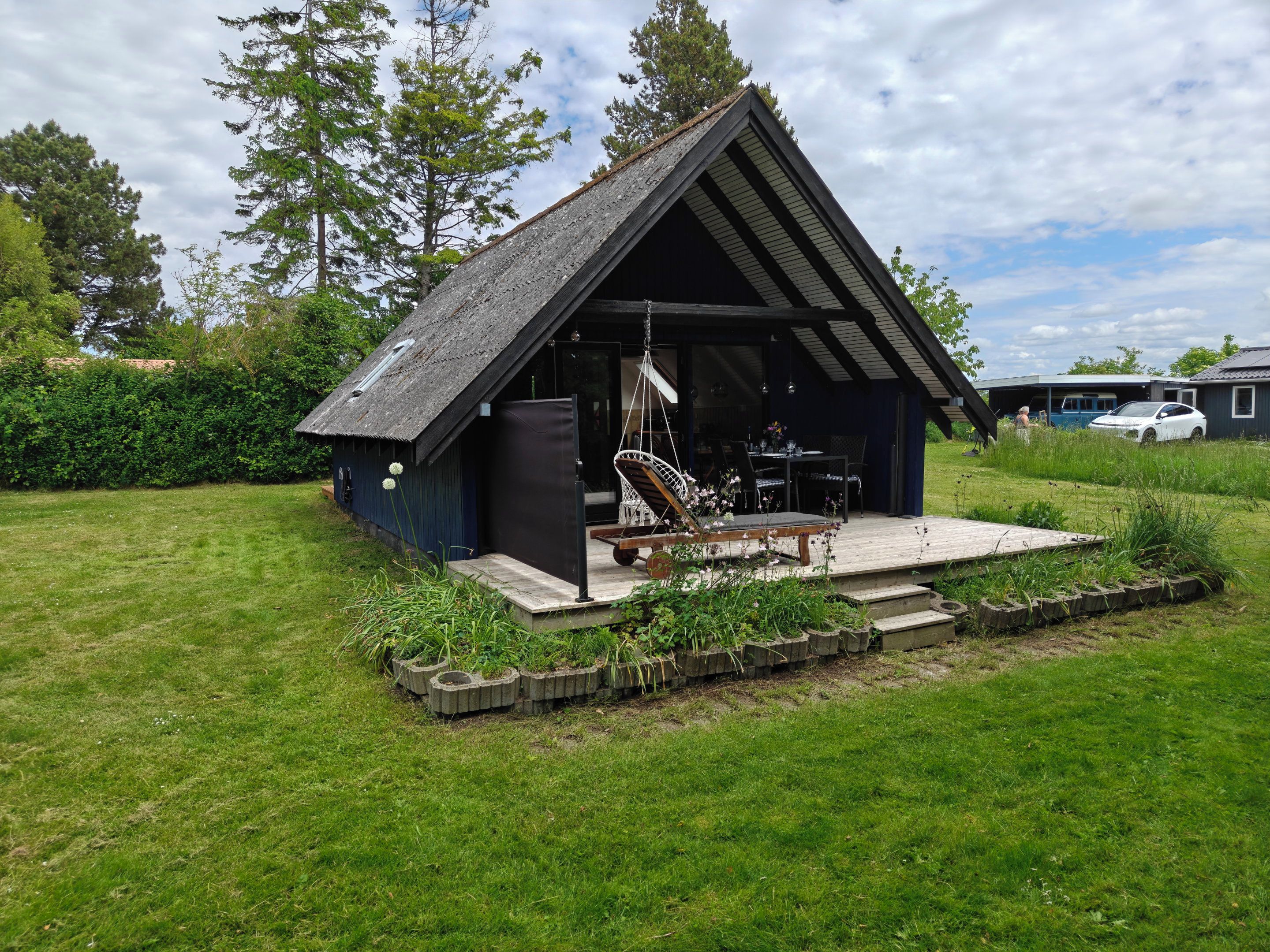 Een klein, donker houten huisje met schuine dak, een terras en een hangstoel.