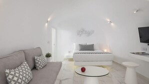 A modern, minimalist living room with a sofa, a round coffee table, a bed with a white headboard, and a TV mounted on the wall. - White Concept Caves - Adults Only (Santorini)