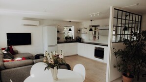 A modern living room with a sofa, a dining table with chairs, a flat-screen TV mounted on the wall, and a kitchen area with cabinets and a countertop.