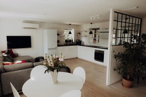 Una sala moderna con un sofá, una mesa de comedor con sillas, un televisor de pantalla plana montado en la pared y una zona de cocina con armarios y una encimera.