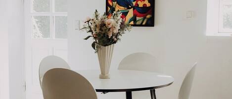 Un comedor con una mesa blanca, cuatro sillas, un florero con flores y un cuadro abstracto colorido en la pared.