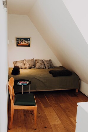 Una habitación pequeña y acogedora con techo inclinado, una cama con ropa de cama estampada, una sola silla, una mesita con libros y un cuadro en la pared.