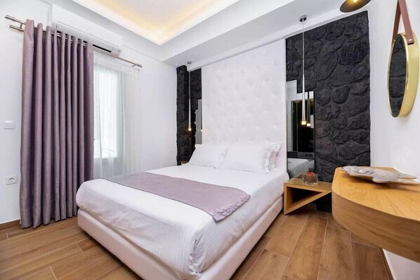 A modern bedroom with a large bed, a wooden bedside table, a stone-textured wall, and a round mirror. - Deluxe Hotel Santorini (Santorini)