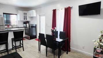 A modern kitchen with white cabinets, a dining area with black chairs, a ceiling fan, and a flat-screen TV mounted on the wall.