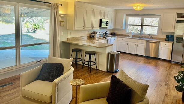 A modern kitchen with white cabinets, a stainless steel refrigerator, and a large window. There are two bar stools and a small round table. - Panther Creek Cozy Retreat (Newton)