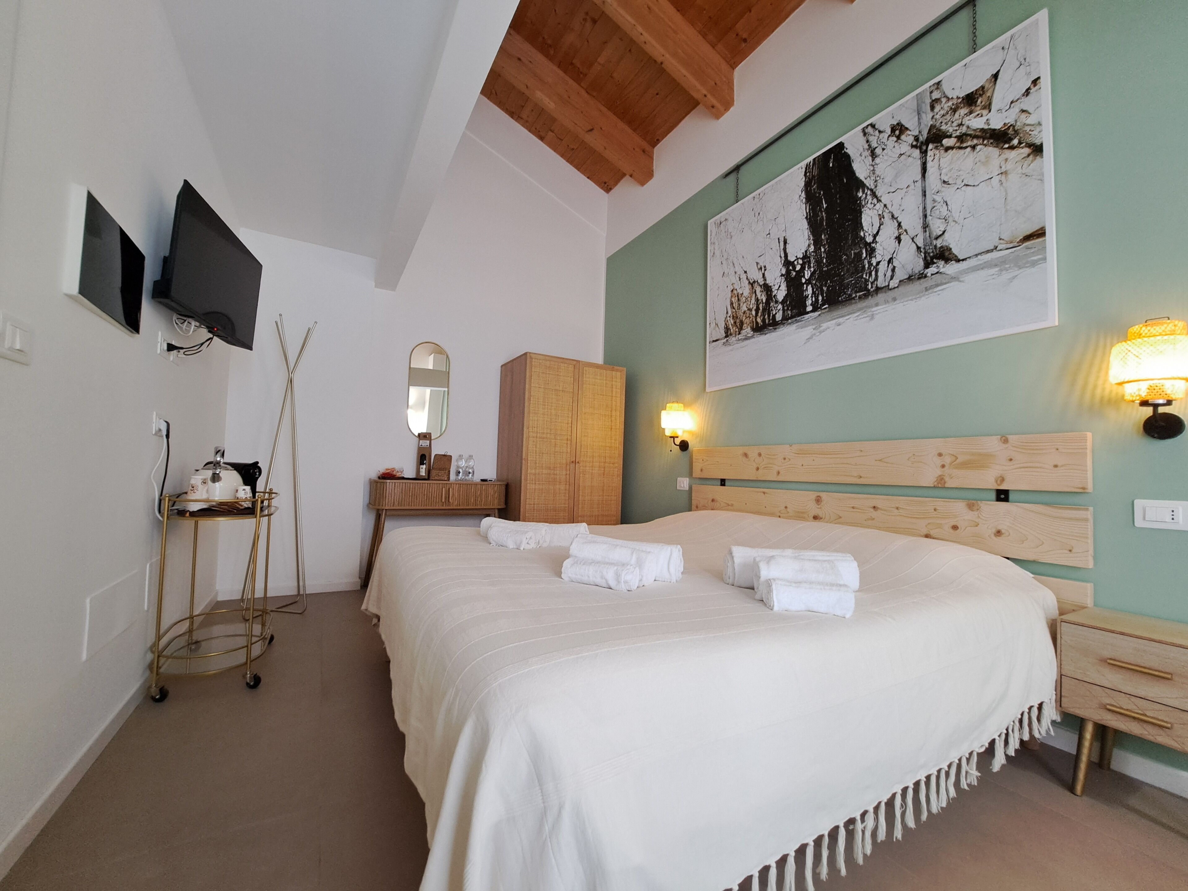 A hotel room with a large bed, a wooden headboard, a wall-mounted TV, a bedside table with a lamp, and a painting on the wall.