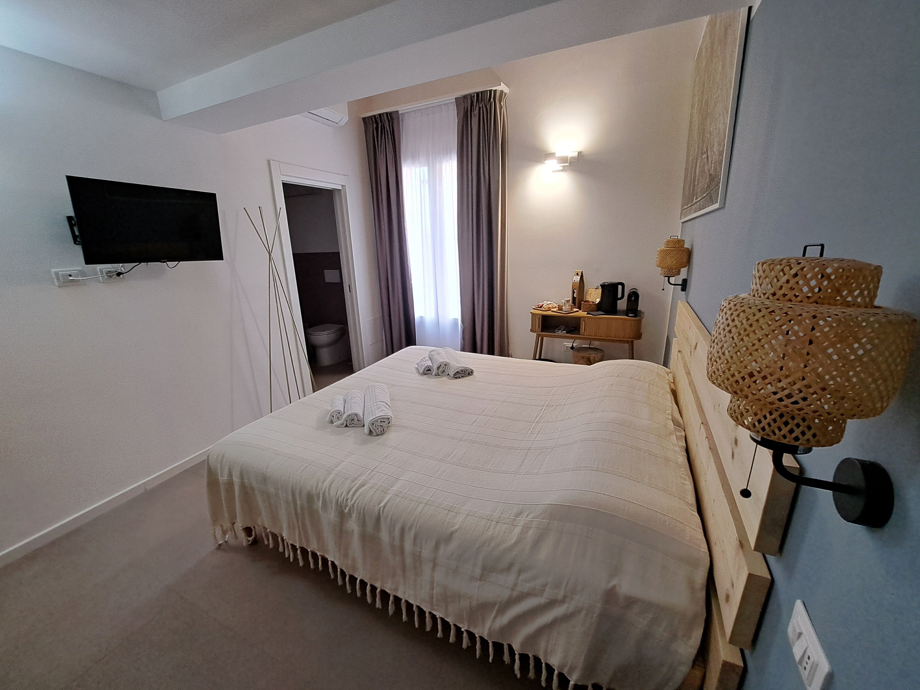 A hotel room with a bed, a television mounted on the wall, a bedside table with a lamp, and a window with curtains.