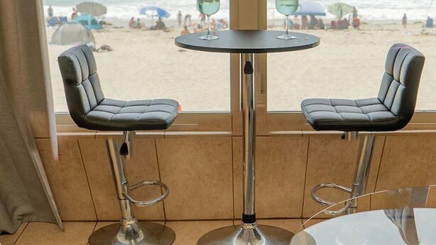 A bar area with two chairs and a table, overlooking a beach with people and umbrellas.