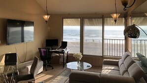 A living room with a large window overlooking the beach, a flat-screen TV, a desk with a computer, and a sofa.