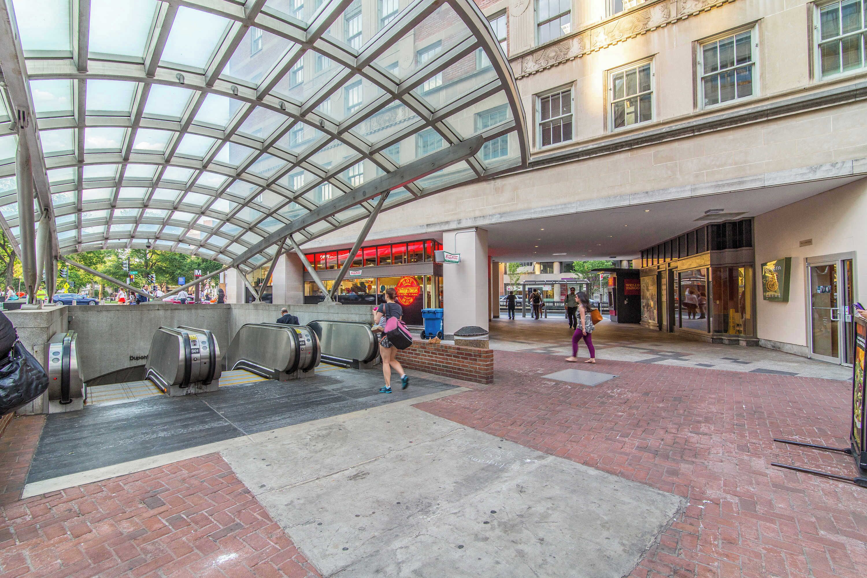 Une zone piétonne abritée, dotée d’escalators et d’une verrière, qui mène à un bâtiment abritant des boutiques.