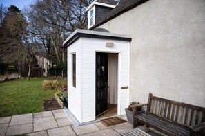 A small, white, single-story structure with a black door, a bench, and a potted plant nearby. - Mill Cottage - a tranquil cottage location for 4 guests (Forres)