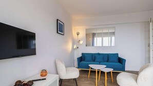 Una sala de estar con una televisión de pantalla plana, un sofá azul, dos sillas blancas, una mesita blanca y un aplique de pared.
