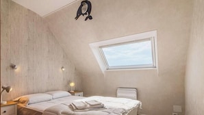 Un dormitorio con techo inclinado, tragaluz, cama con ropa blanca y una lámpara fijada a la pared.