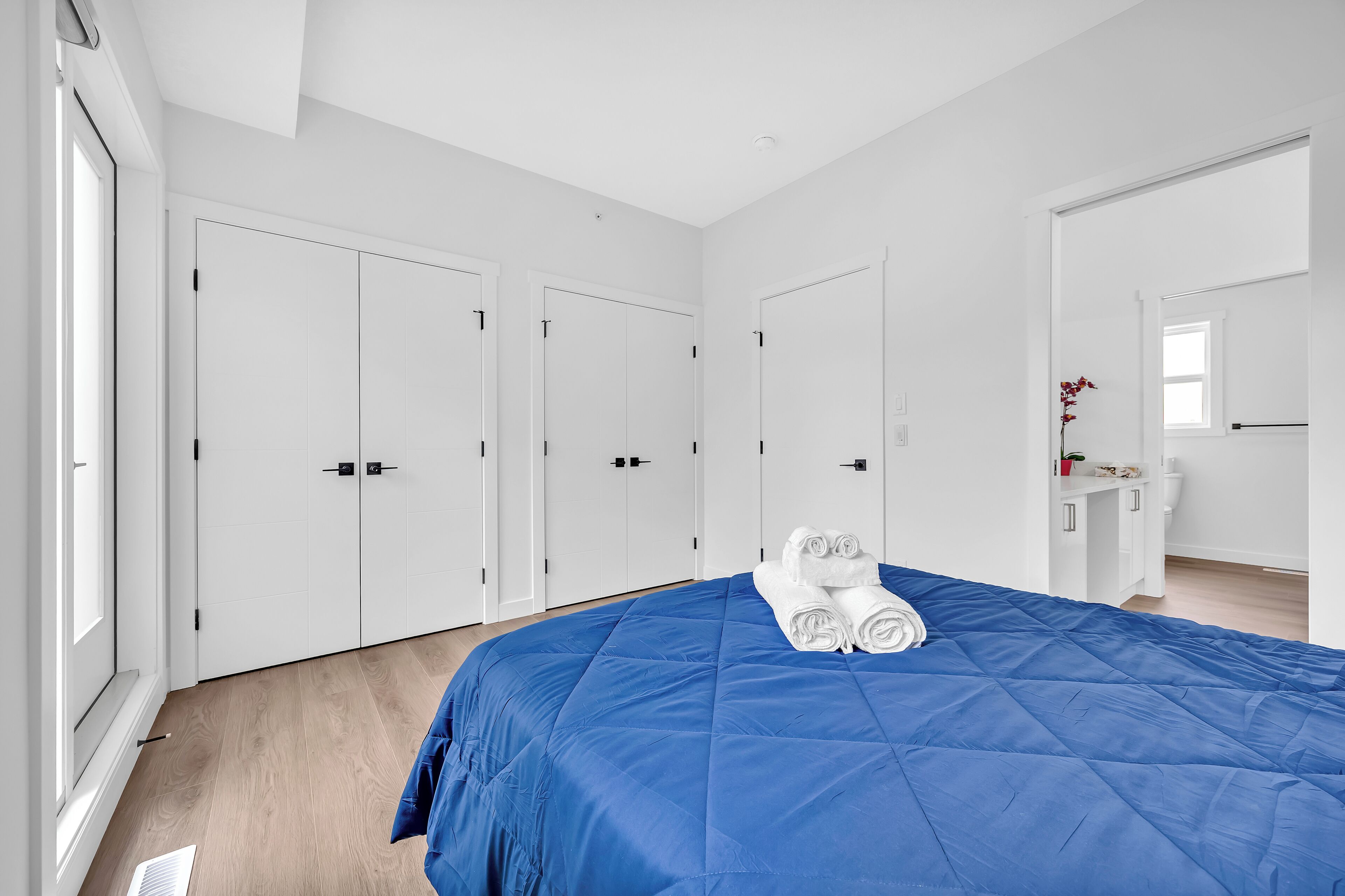 A bedroom with a blue bedspread, white wardrobe, and a small bedside table with a red flower.