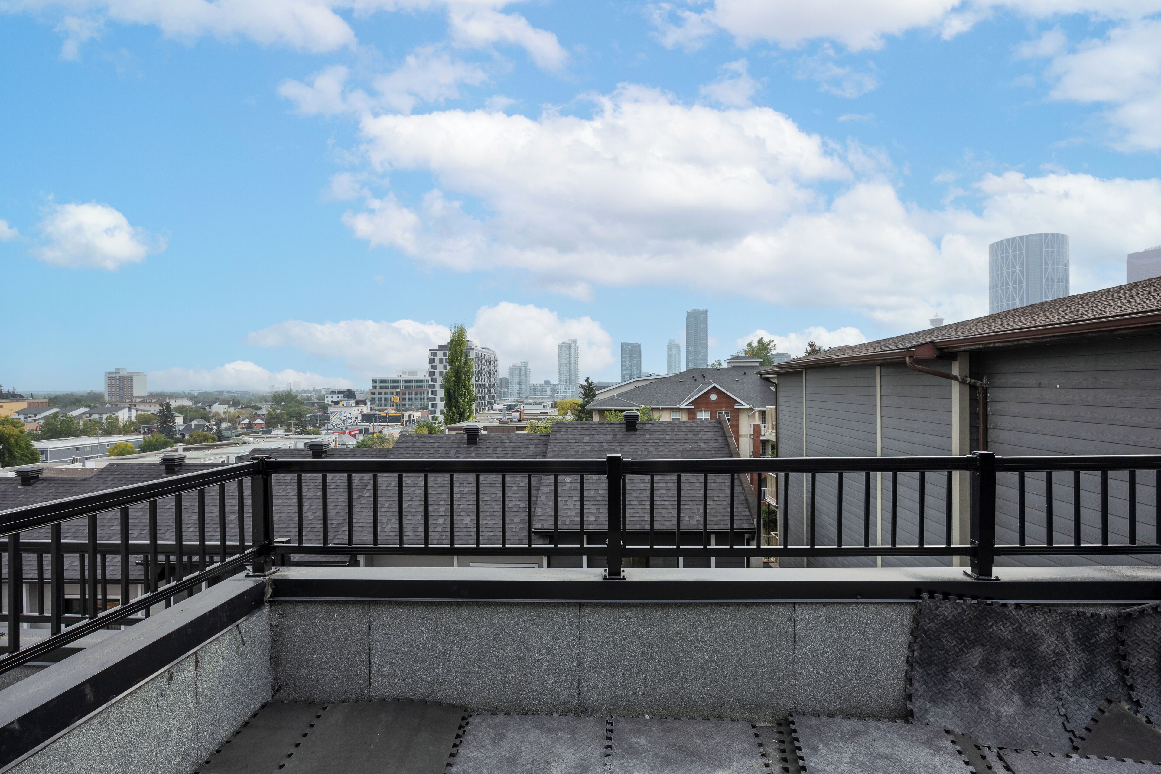 Una terraza con barandilla negra, piso de concreto y vista de un paisaje urbano con edificios y cielo despejado.