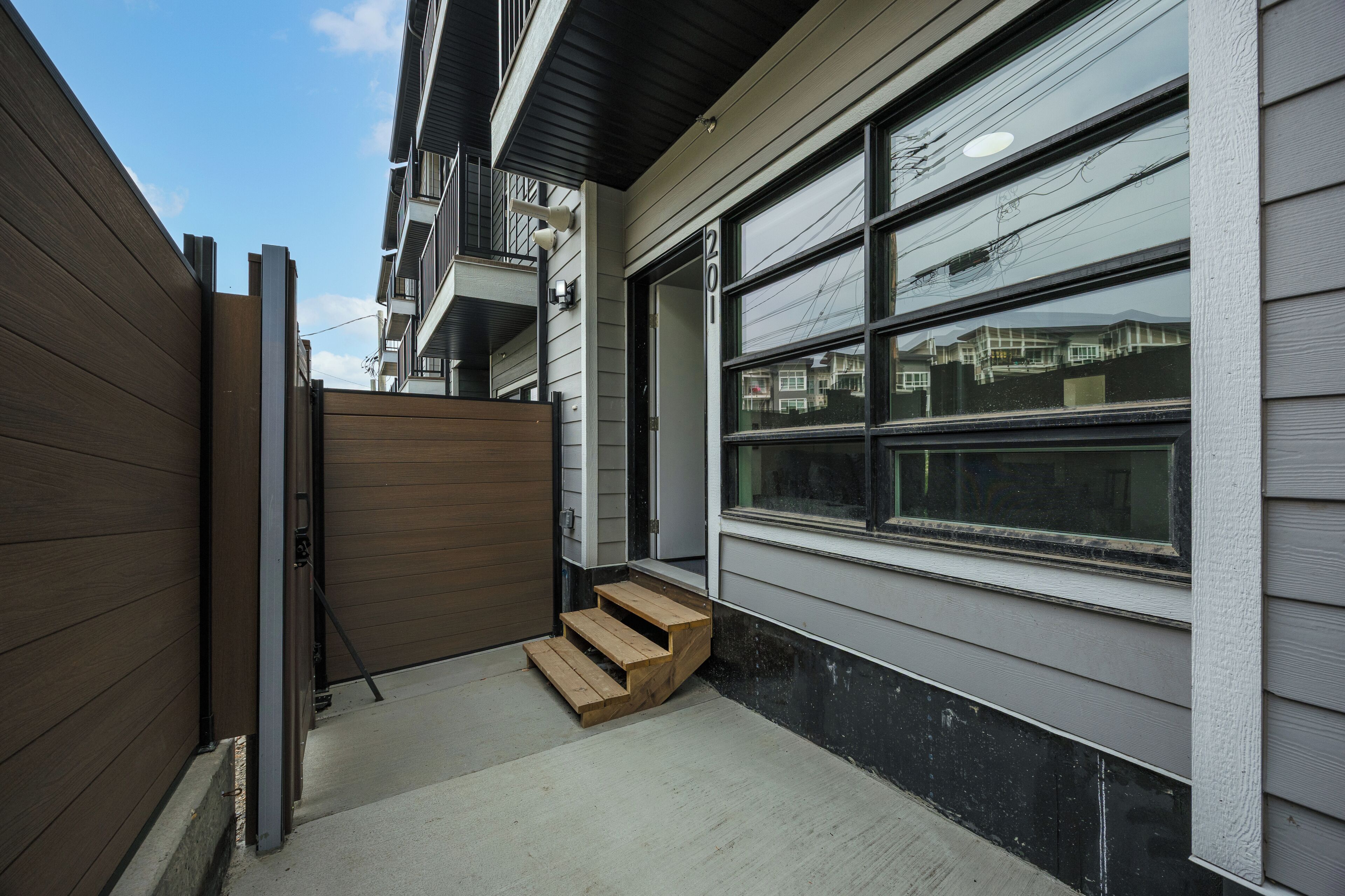 Un edificio residencial con un portón de madera, una escalera de madera que lleva a una puerta y grandes ventanales que reflejan edificios vecinos.