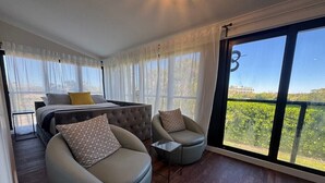 A modern bedroom with a bed, two armchairs, a small table, and large windows offering a view of greenery and buildings.